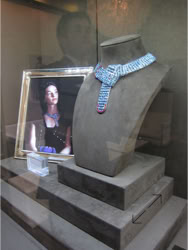Basel Display Case A display case containing a framed portrait of a person and a bejeweled necklace on a stand.