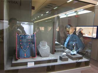 Basel Display Case A display case with various jewelry items, including a beaded necklace on a stand, a pearl necklace, and other small items. A person is looking at the display through the glass Basel Display Case A decorative clock with a round face and ornate base displayed on a pedestal, with other objects in the background design of stand and displays by Swiss Packaging of New York and Switzerland
