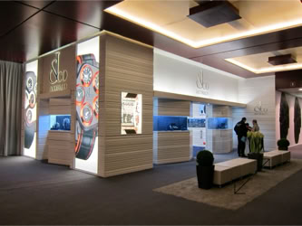 Basel Fair jacob & Co Exhibition booth with illuminated signage, large watch images, and people standing near counters design and design by Swiss Packaging of New York and Switzerland
