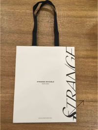 A white shopping bag with black handles, featuring the word "STRANGE" vertically on the right side and Strange Invisible text in the center created by Swiss Packaging