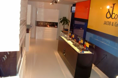A Basel fair exhibition stand interior with a glossy floor, a display counter with items, a large colorful wall with Jacob & Co. logo, and a potted plant in the background design and design by Swiss Packaging of New York and Switzerland