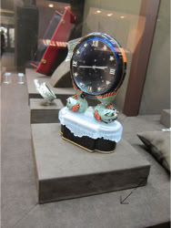 Basel Display Case A decorative clock with a round face and ornate base displayed on a pedestal, with other objects in the background design of stand and displays by Swiss Packaging of New York and Switzerland