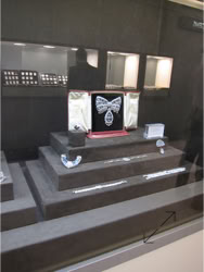 Basel Display Case A display case with a tiered platform showcasing jewelry, including a large butterfly-shaped piece in the center, surrounded by smaller jewelry items.