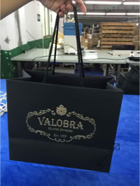a black shopping bag with gold decorative text and design, reading "VALOBRA" and "Maison Fondée en 1905," created by Swiss Packaging
