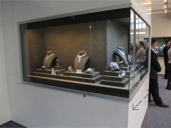 Basel Display Case A glass display case containing various pieces of jewelry, including necklaces and rings, with people visible in the background design of the trade show booth and displays by Swiss Packaging of New York and Switzerland