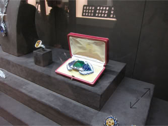 Basel Display Case A display case with a large gemstone set in a red open box, surrounded by other jewelry pieces on a dark surface.