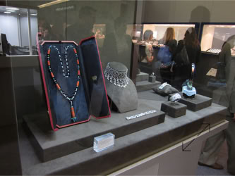 Basel Display Case A display case with various pieces of jewelry, including necklaces and bracelets, set on stands. People are visible in the background Basel Display Case A decorative clock with a round face and ornate base displayed on a pedestal, with other objects in the background design of stand and displays by Swiss Packaging of New York and Switzerland