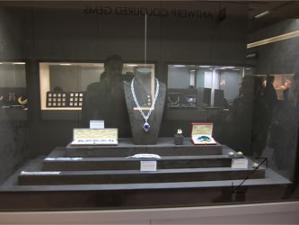 Basel Display Case A display case containing a necklace with a large pendant, several smaller items, and informational cards, with reflections of people in the glass.
